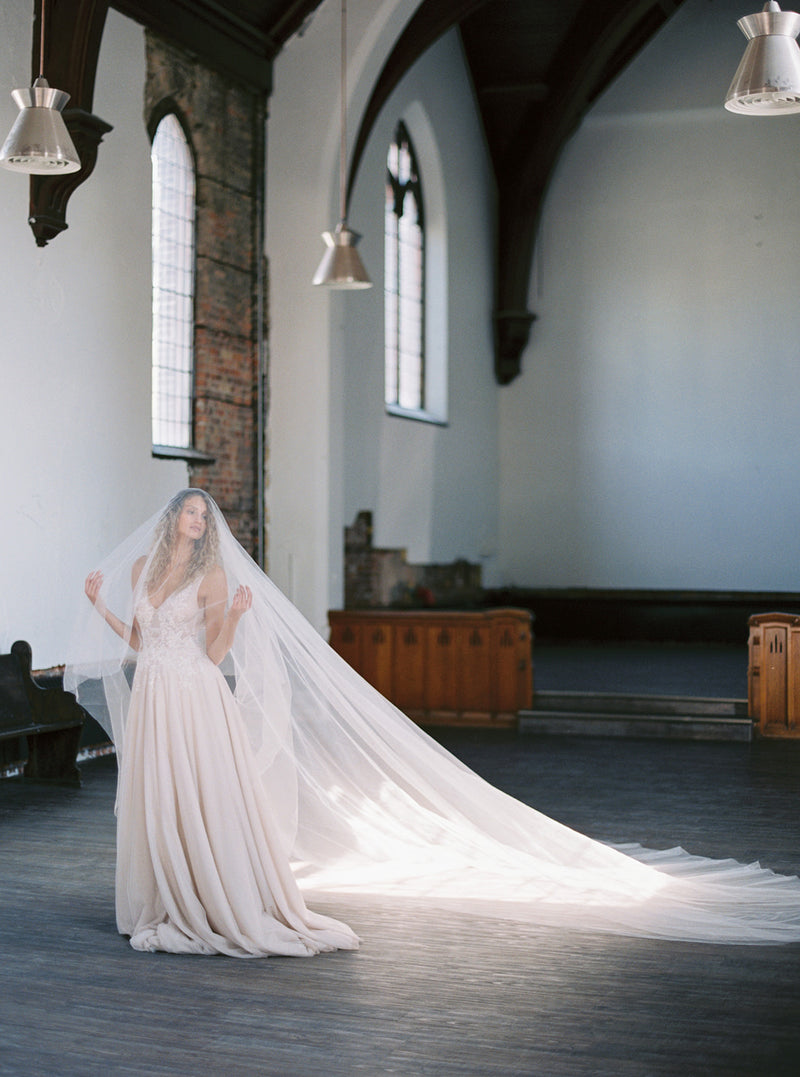 Wide shot of the bride standing alone in a large, bright church, showing the full, dramatic length of the Grace silk wedding veil train laid out.