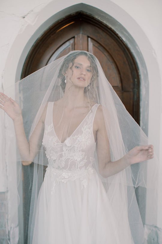 A bride wearing a silk blusher veil and a floral wedding gown is standing in front of an arch church door.