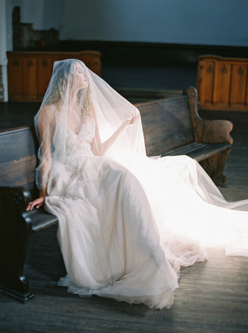 Timeless bridal portrait: Bride in Grace English Silk Veil sitting on an antique church pew, showing the ethereal nature of the tulle.
