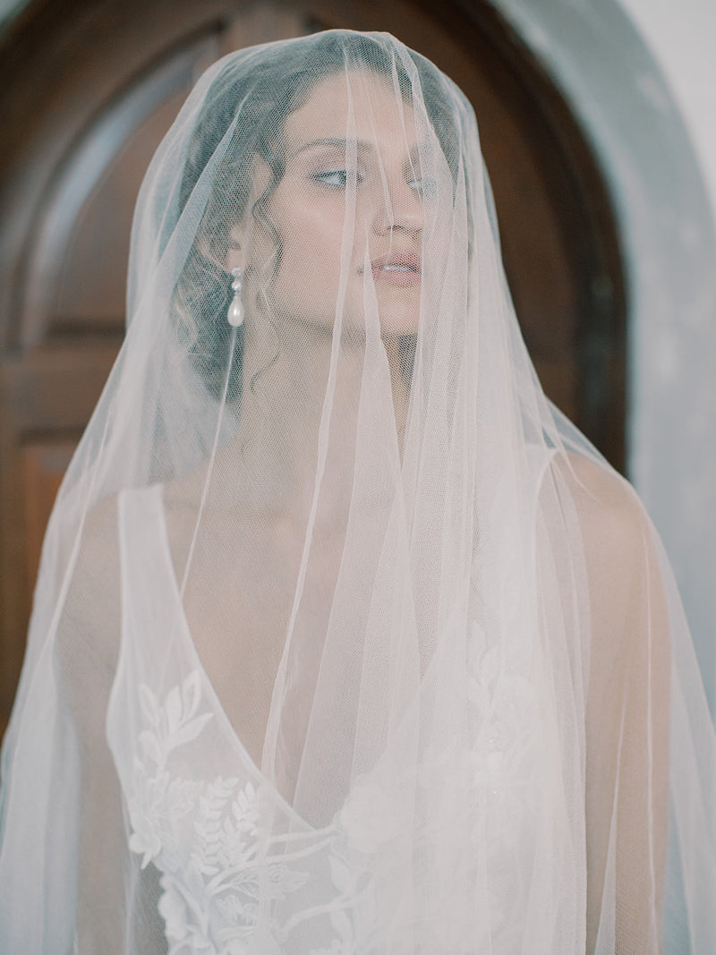 Close portrait of the bride with a soft updo,wearing the Grace silk blusher veil over her face, head slightly turned, clearly showing the fabric's sheer quality and her expression.