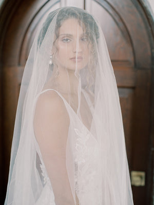 Very close portrait of the bride, the Grace English silk veil covering her face, showing a pearl earring and the slight texture of the silk tulle.