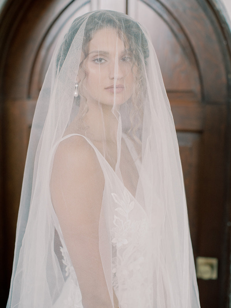 Very close portrait of the bride, the Grace English silk veil covering her face, showing a pearl earring and the slight texture of the silk tulle.