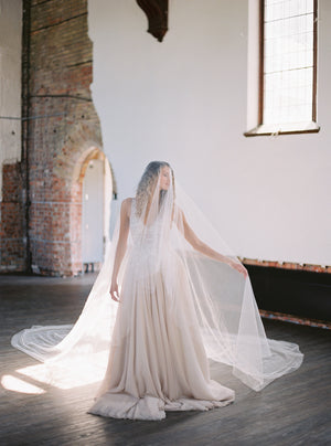 Bride standing in a historic church with the Grace Silk Veil draped over her; the wide veil train is fanned out on the wood floor. 