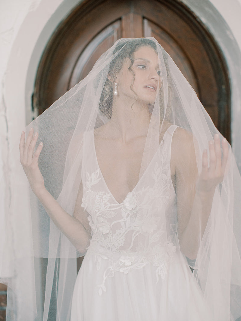 Close-up of the bride framed by the Grace silk blusher veil, hands raised to show the blusher layer pulled forward, with a dark wooden door backdrop.