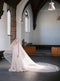 Full view of the bride standing in a church with arched windows, the Grace Silk Blusher Veil train is laid out on the wood floor, showcasing the majestic width and ethereal transparency of the English silk tulle.