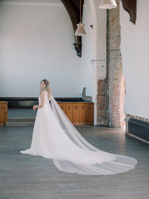 KAJA draped bridal cape veil styled with a lace gown and half up hairstyle. The long train is beautifully fanned out behind the bride.