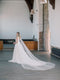 KAJA draped bridal cape veil styled with a lace gown and half up hairstyle. The long train is beautifully fanned out behind the bride.