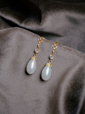 A pair of LYON bridal drop earrings featuring a gold setting, clear crystals, and an oval teardrop pearl resting on textured velvet fabric.