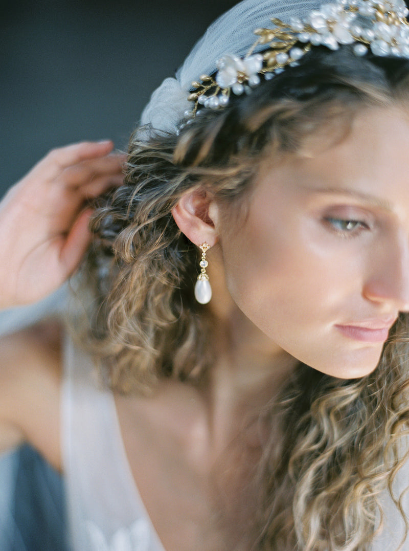 Close-up of a bride wearing the LYON pearl and crystal drop earrings and a pearl wedding headband and Juliet cap veil. The earring's crystal stud and pearl drop are clearly visible against her hair.