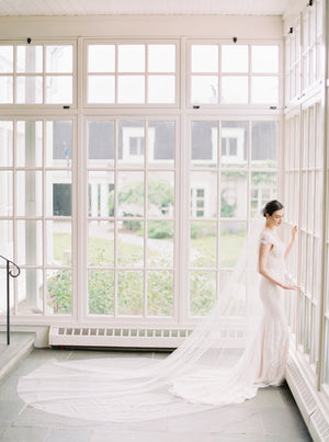 A bride in a lace gown standing in the sunroom at Langdon Hall, with the Olivia long wedding veil fanned out dramatically on the slate floor behind her.