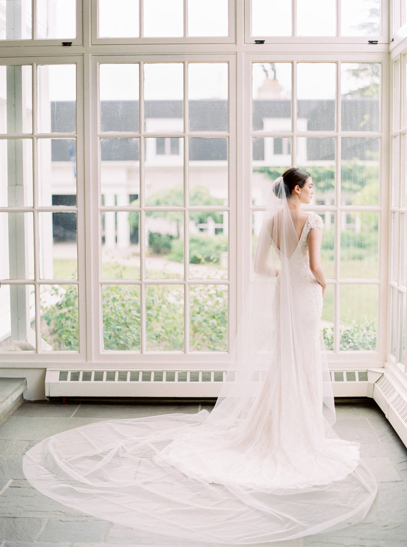 Back view of the bride wearing a fitted lace gown and the Olivia wedding veil. The wide train of the veil is fanned out on the ground behind the bride.