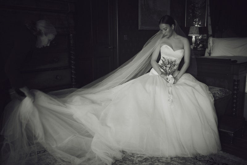 Intimate moment before a wedding, showing the flowing, sheer Olivia veil draped over a bride holding a small bouquet while sitting on a four-poster bed.