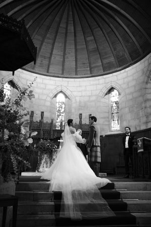Black and white photo of a bride and groom during their ceremony; the bride's extra-long Olivia wedding veil flows dramatically down the steps of the stone church altar.