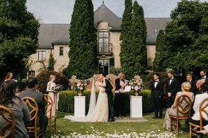 Bride and groom kissing at the altar during an outdoor wedding ceremony, featuring the Olivia long veil trailing majestically across the petal-strewn grass.
