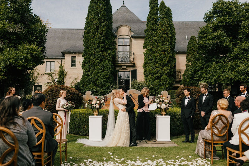 Bride and groom kissing at the altar during an outdoor wedding ceremony, featuring the Olivia long veil trailing majestically across the petal-strewn grass.