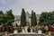 Wide shot of an elegant outdoor wedding ceremony; the long Olivia veil is visible behind the bride, who is standing under two tall trees with a stately manor in the background.