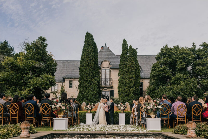 Wide shot of an elegant outdoor wedding ceremony; the long Olivia veil is visible behind the bride, who is standing under two tall trees with a stately manor in the background.
