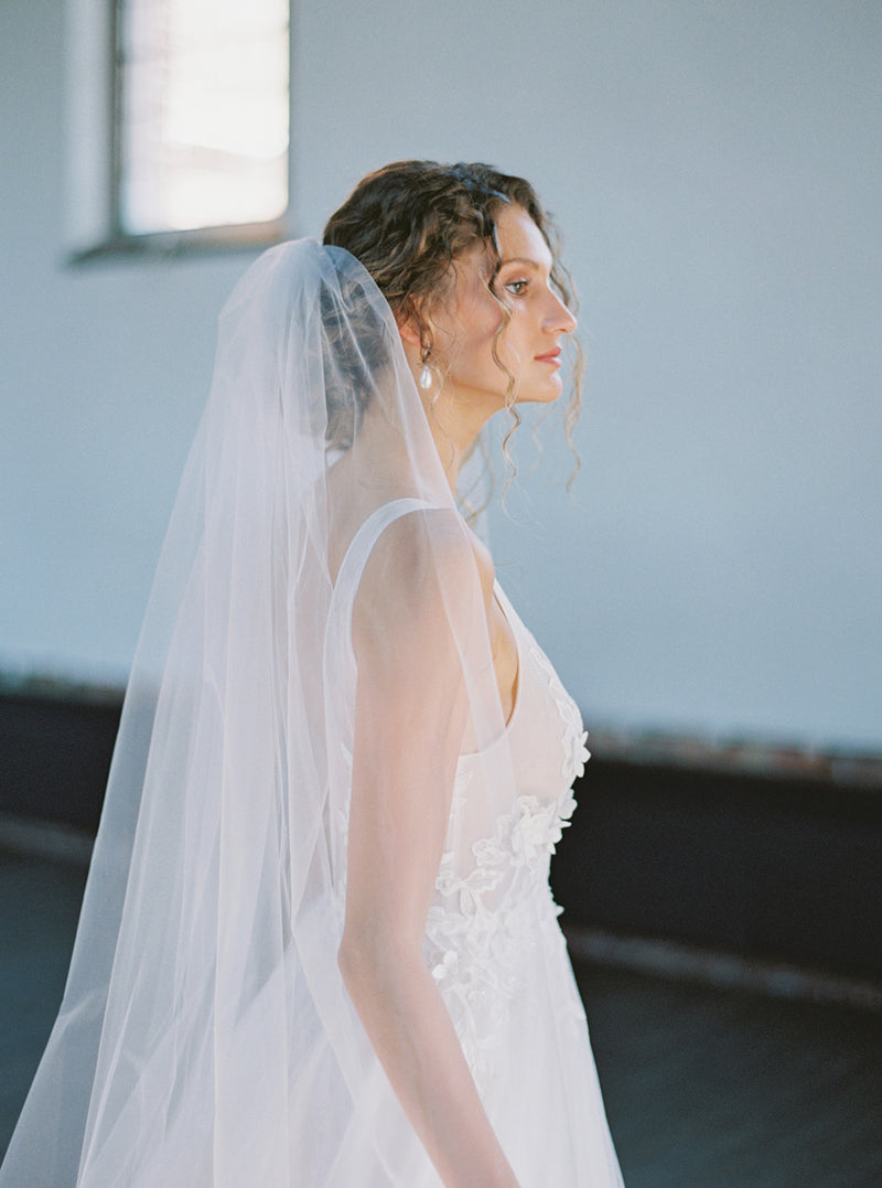 Bride in side profile wearing the Rosanna full wedding veil. The fully gathered tulle is visible at the comb, creating a voluminous layer that flows behind her shoulders.