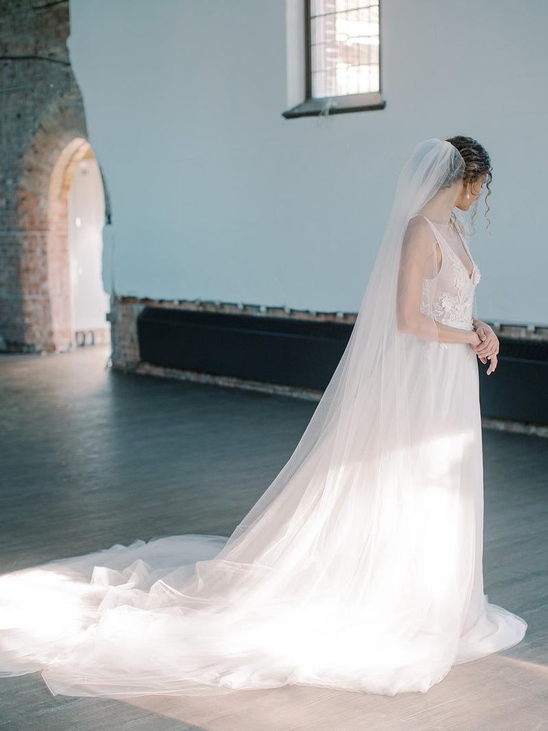Side profile of a bride in the Rosanna full wedding veil. The veil's wide train is fully fanned out on the wood floor.