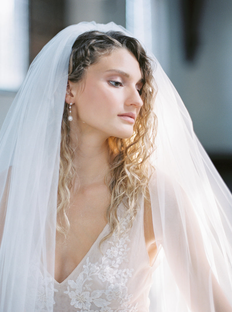 Close-up bridal portrait showing the full Rosanna wedding veil draped over the bride's curly hair, highlighting the sheer, voluminous Whisper tulle and her pearl earrings.