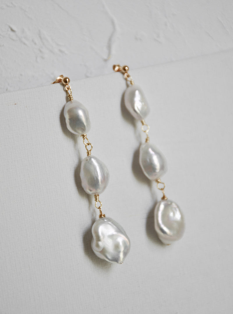 Pair of Tia long drop wedding earrings, showing the graduating size of the baroque freshwater pearls against a textured white background.